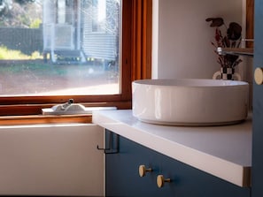 Une salle de bain avec un lavabo rond blanc, une fenêtre donnant sur une maison et un meuble avec un porte-serviettes.