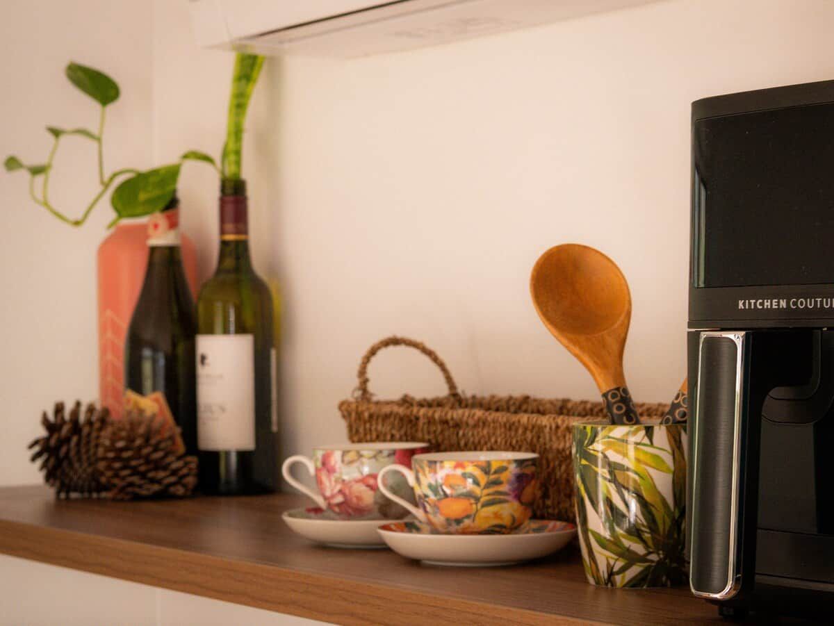 A kitchen counter with a coffee maker, a wicker basket, a wooden spoon, and decorative items including a pine cone and a plant.