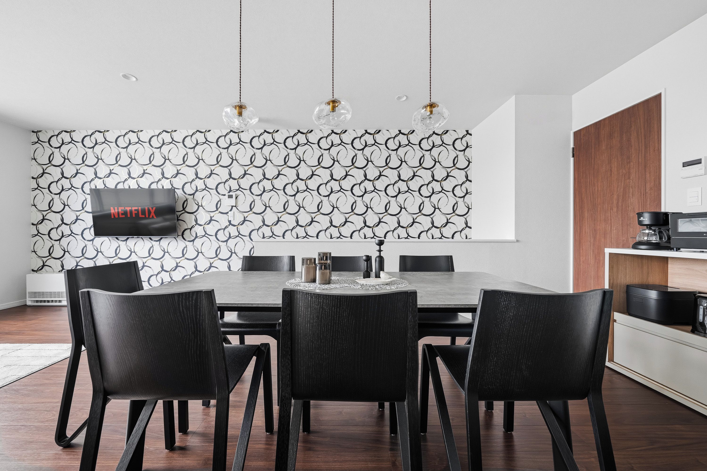 Un comedor moderno con una pared de diseño geométrico, un televisor de pantalla plana y una cafetera.