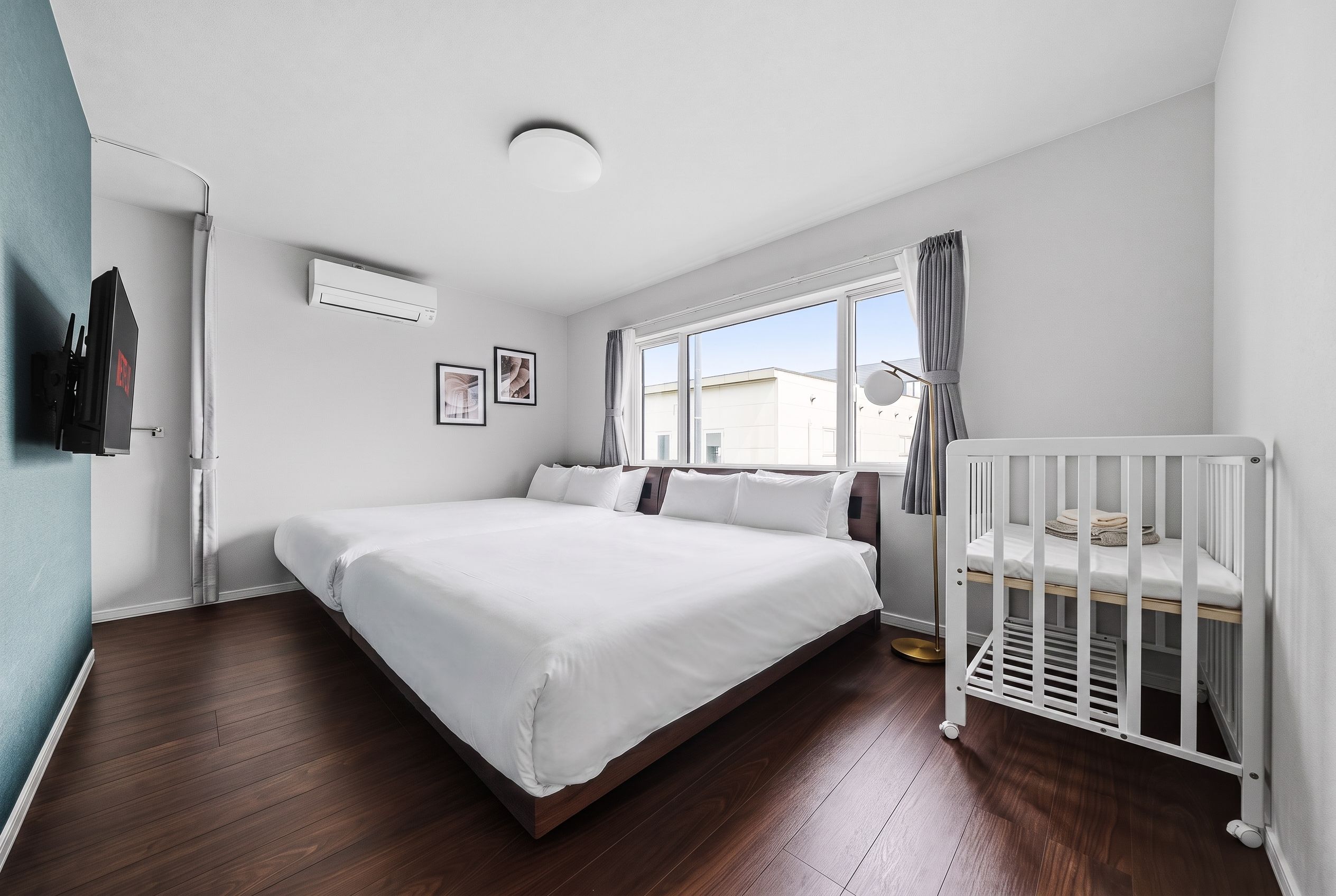 Un dormitorio moderno con una cama grande, una cuna, una ventana con cortinas, un aire acondicionado de pared y una puerta con perchero.