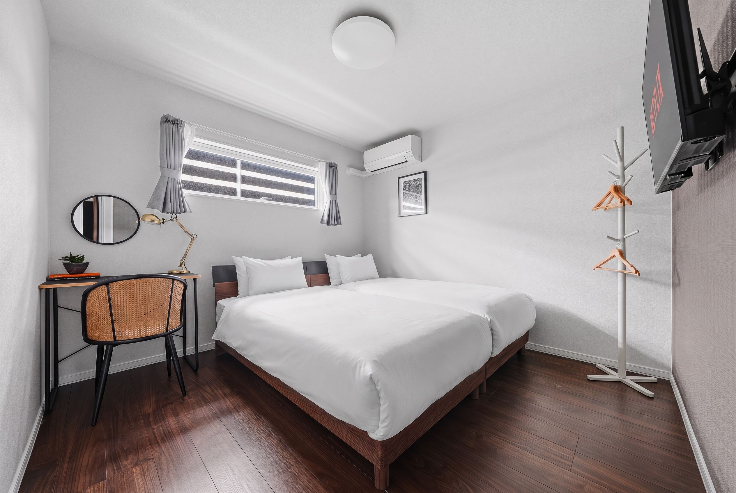 A hotel room with a large bed, a desk with a chair, a window with curtains, a wall-mounted air conditioner, and a coat rack.