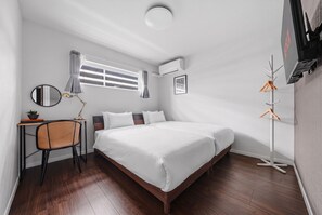 A hotel room with a large bed, a desk with a chair, a window with curtains, a wall-mounted air conditioner, and a coat rack. - Flower (Furano)