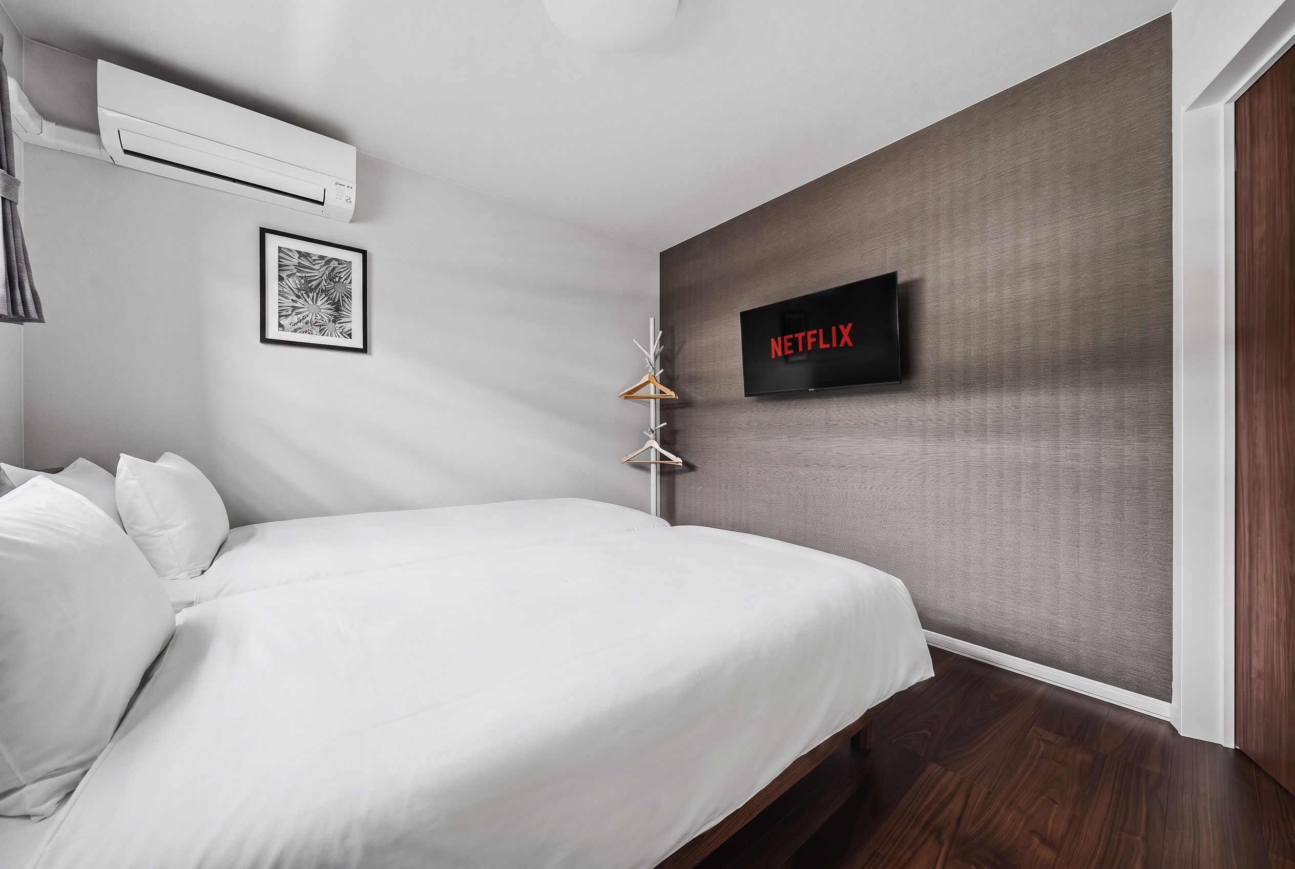 A modern bedroom with a bed, an air conditioning unit, a framed picture, a wall-mounted television, and a wooden floor.