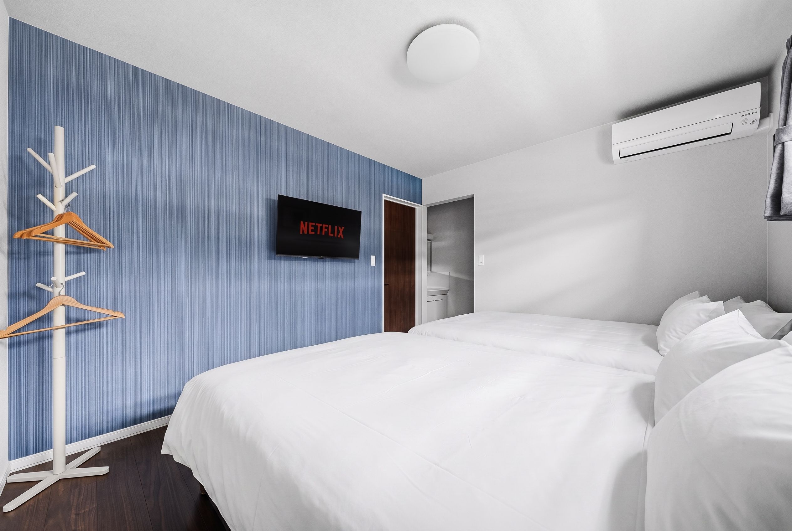 A hotel room with a bed, a TV mounted on the wall, a coat rack, and an air conditioning unit.
