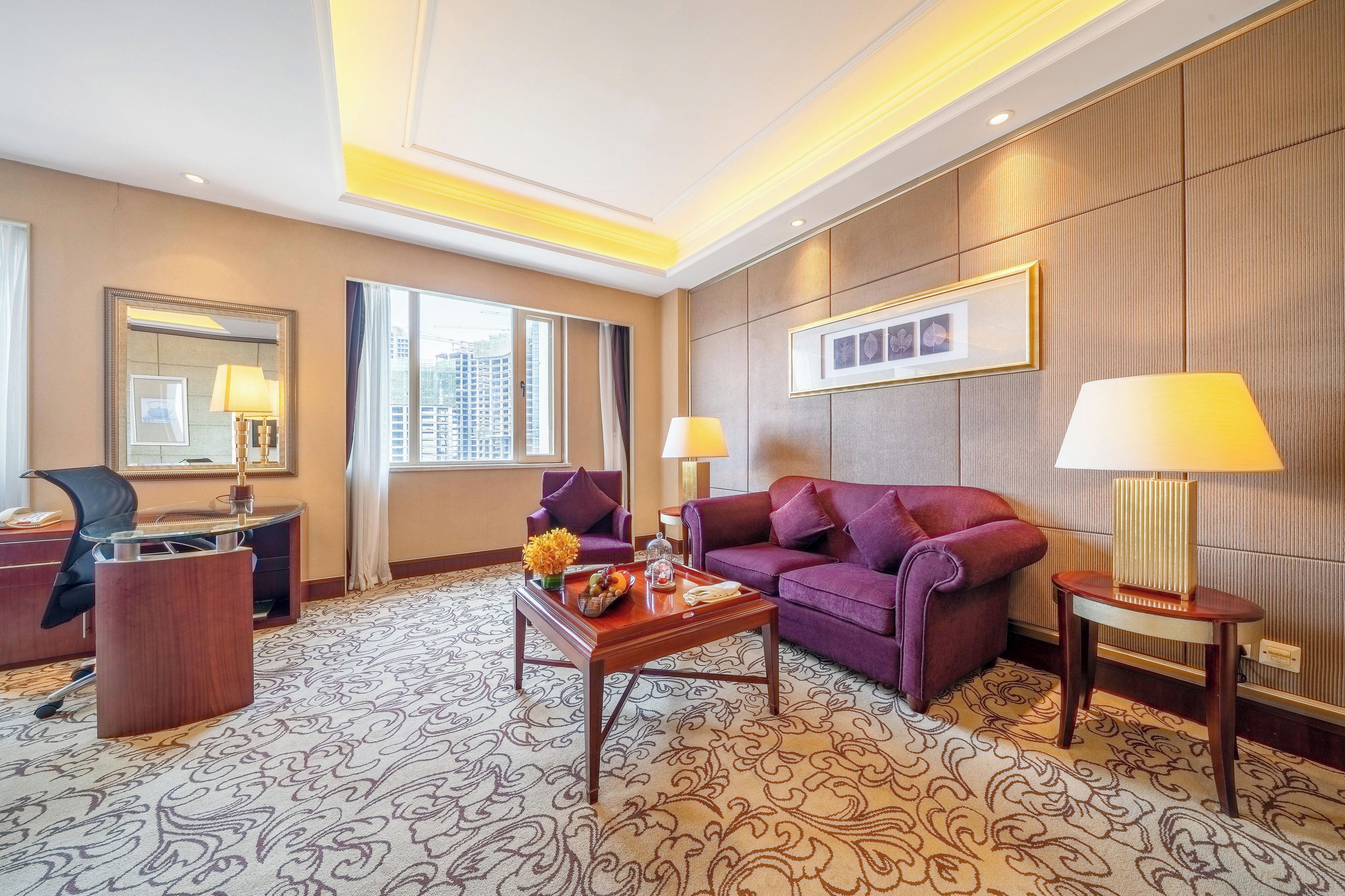 A hotel room with a sofa, armchairs, a desk, a lamp, a mirror, and a patterned carpet.