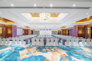 Un salón de conferencias con filas de mesas y sillas, una lámpara de araña grande y una alfombra estampada.