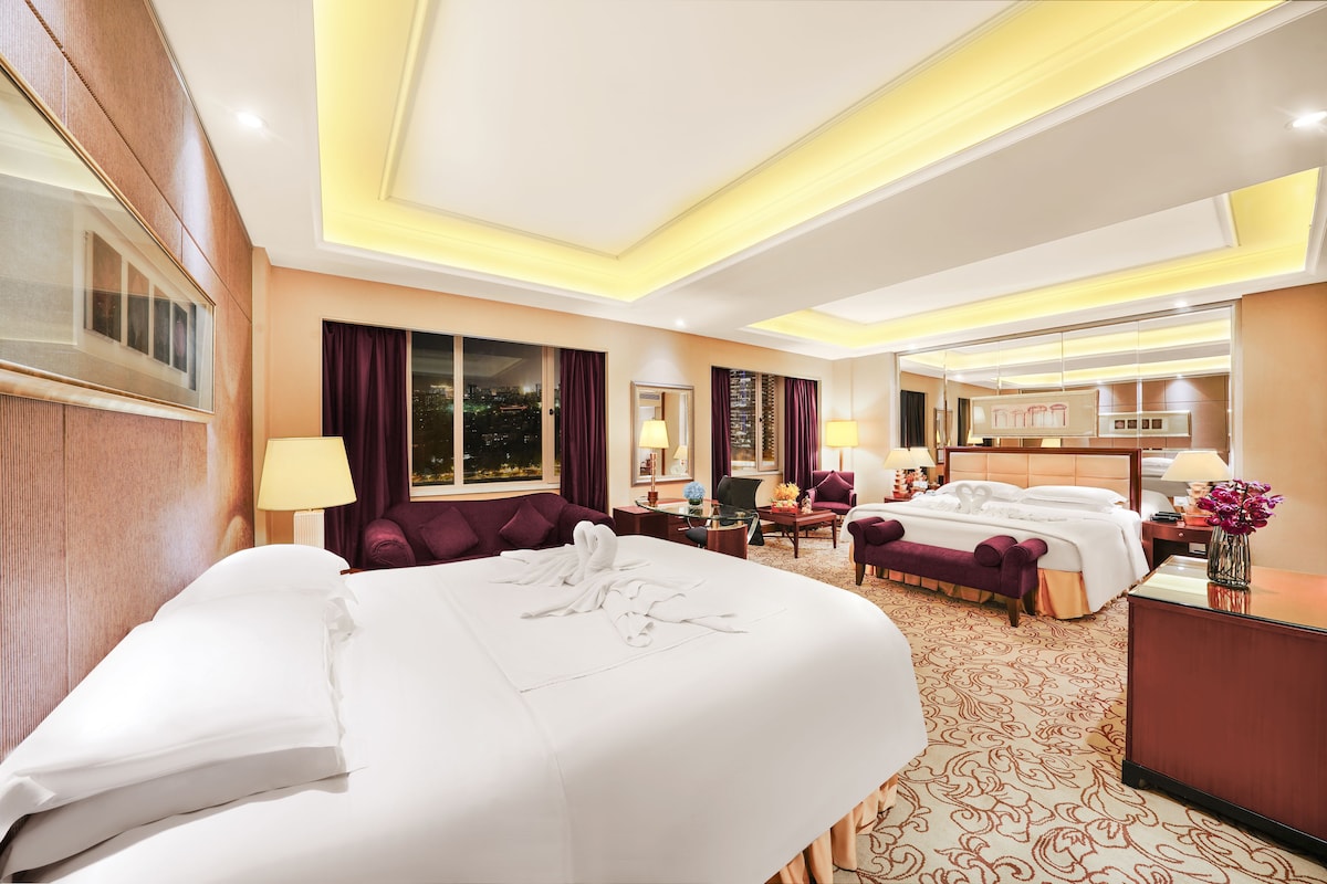 Habitación de hotel con dos camas, una zona de estar y vista a la ciudad de noche.