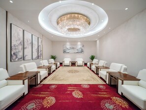 Un salón de conferencias con una mesa redonda, sillas blancas, alfombra roja y un candelabro.