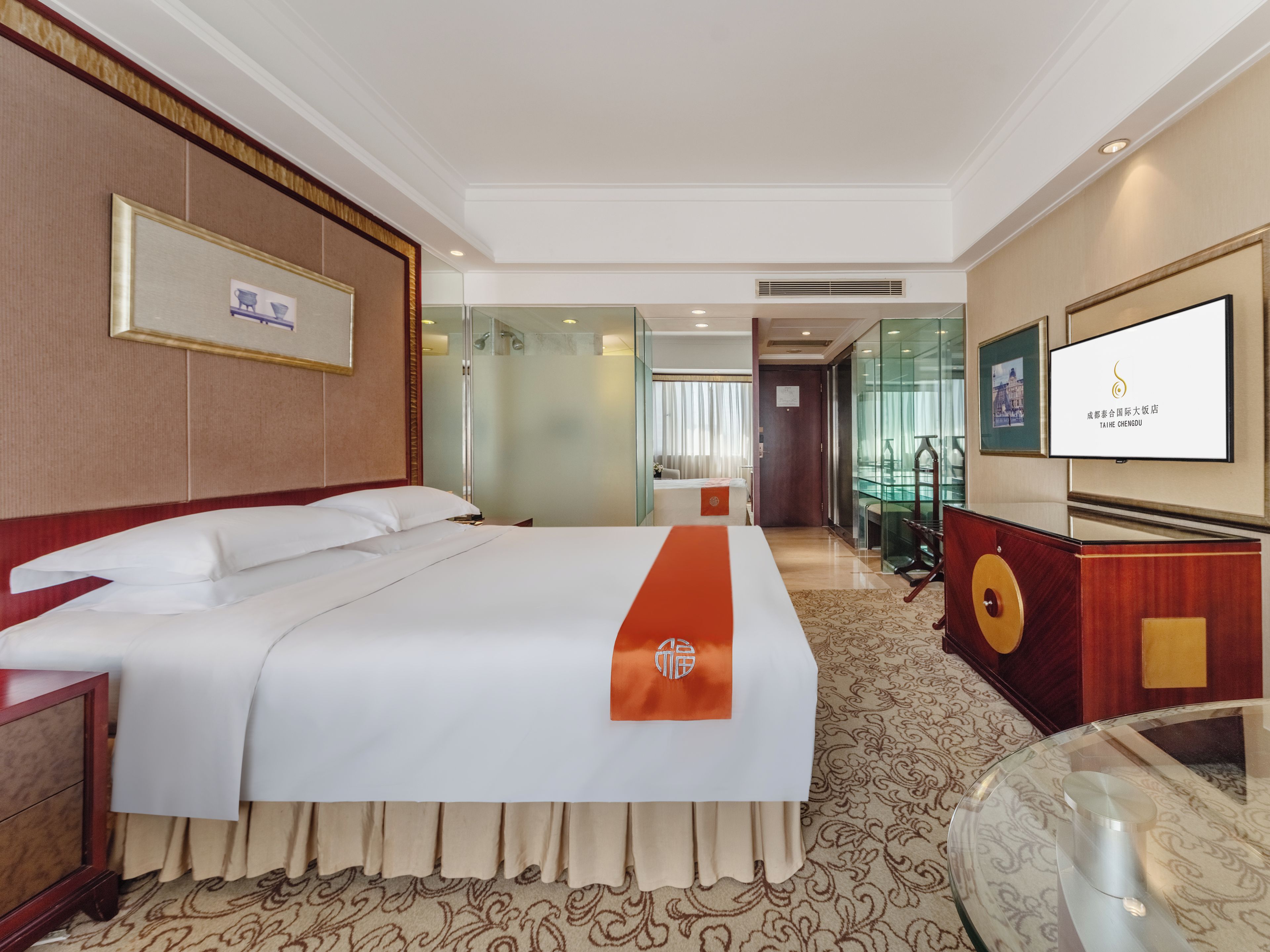 A hotel room with a large bed, a desk, a television, and a bathroom visible through a glass partition.