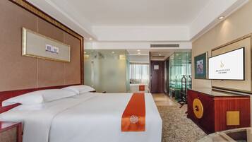 A hotel room with a large bed, a desk, a television, and a bathroom visible through a glass partition.