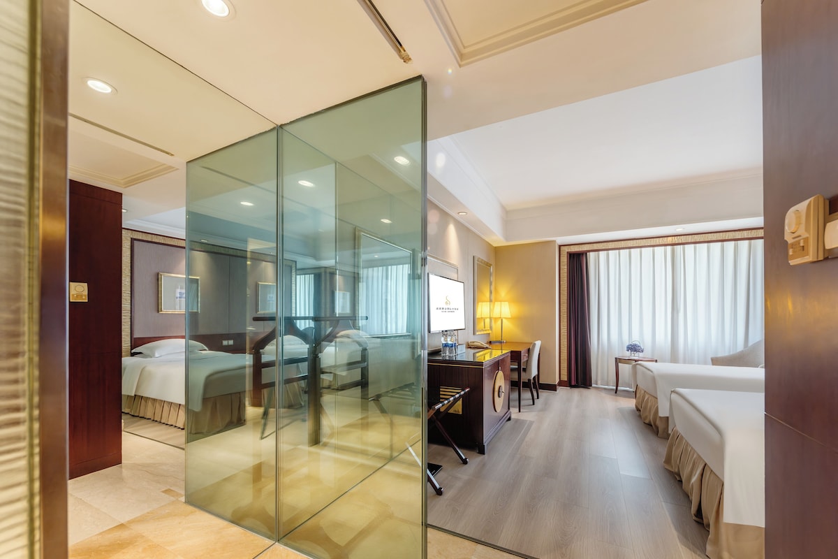 Habitación de hotel con separador de vidrio, cama, escritorio con silla y una mesita de noche con lámpara.