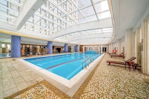 Una piscina interior con techo de vidrio transparente, suelo de baldosas y sillones de descanso.