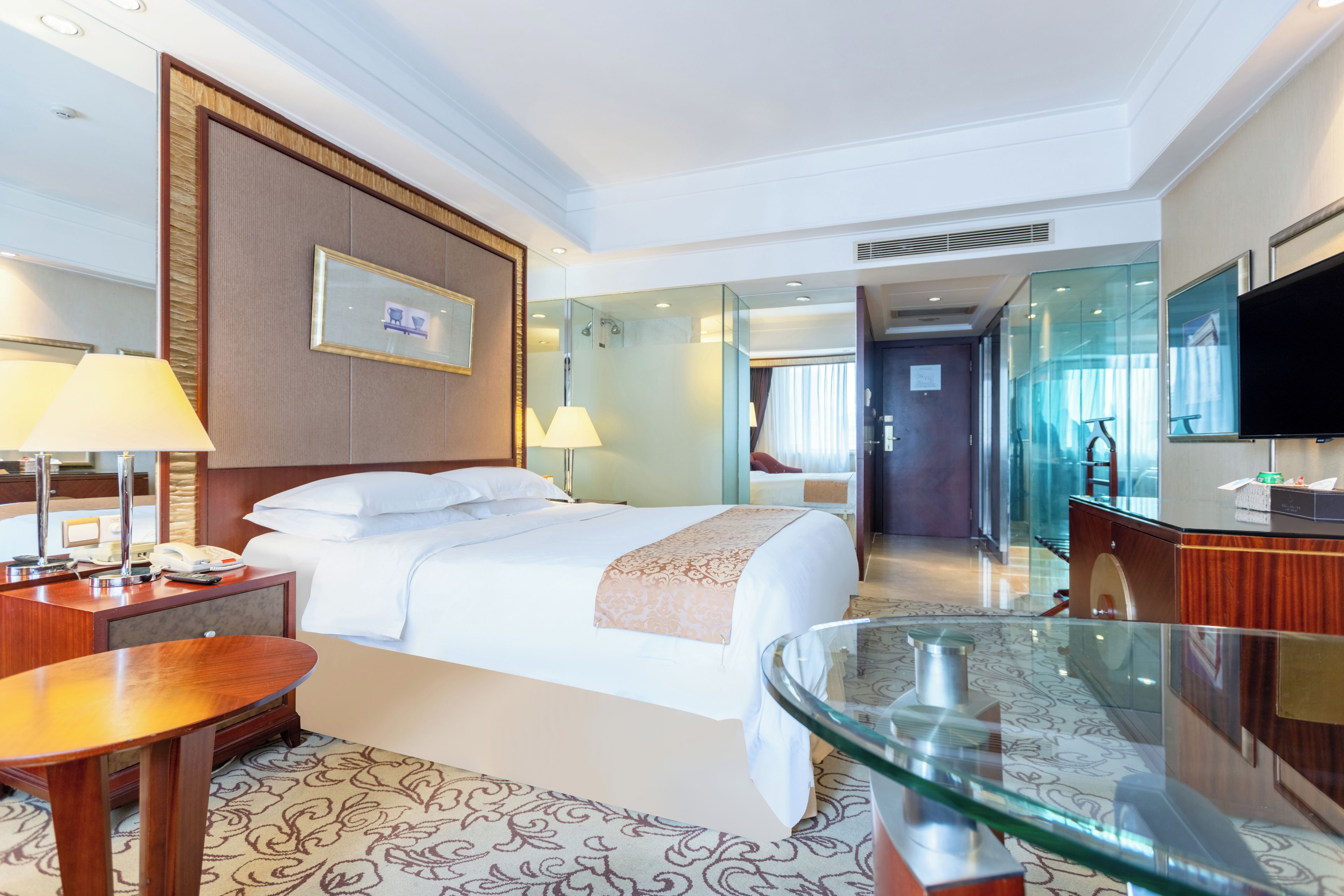 A hotel room with a bed, bedside tables, a desk, a glass coffee table, a television, and a bathroom visible through a glass partition.