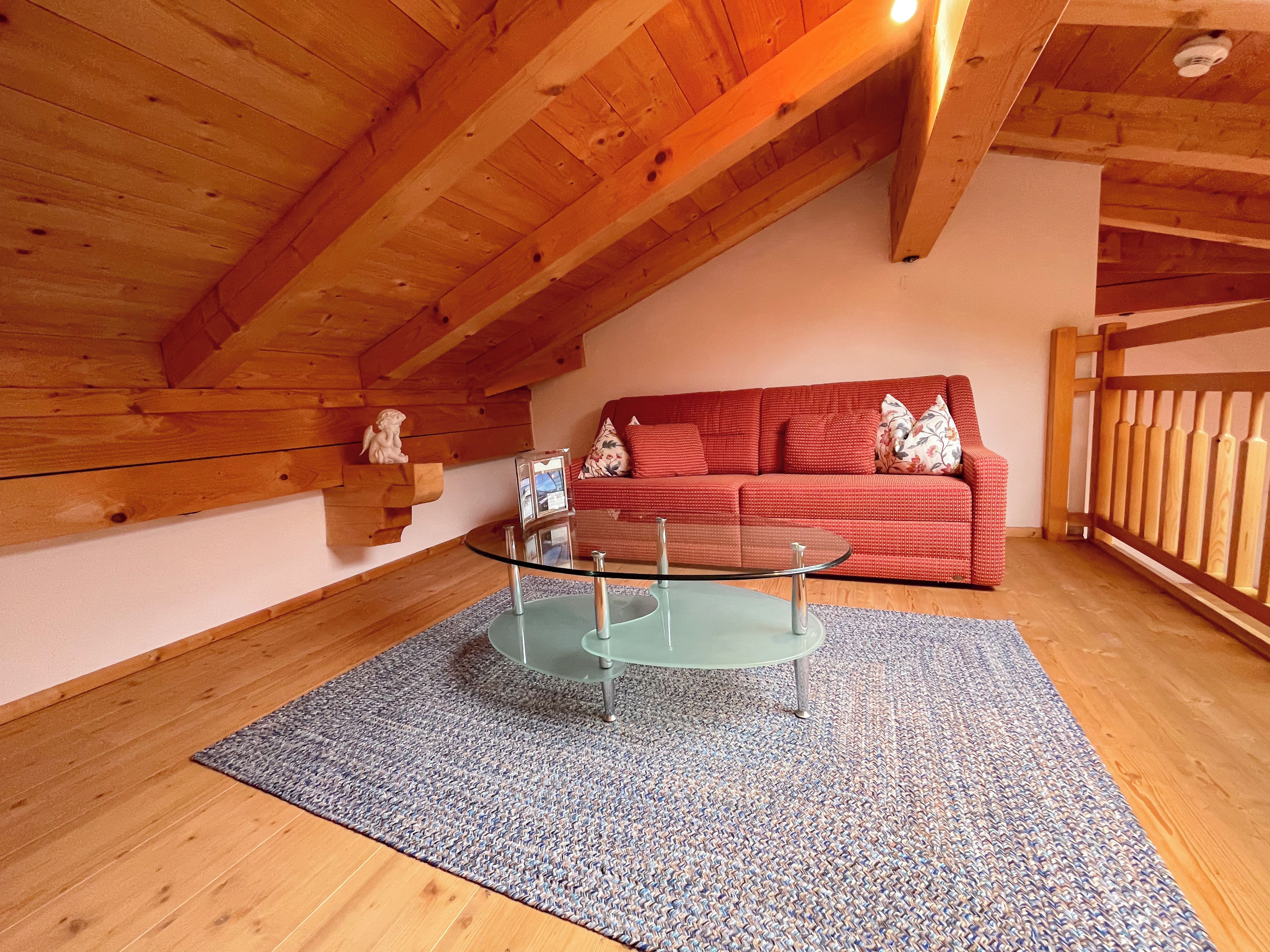 Ein gemütliches Zimmer mit Holzbalkendecke, einem Glastisch, einem roten Sofa und einem kleinen Regal mit einer Statue.