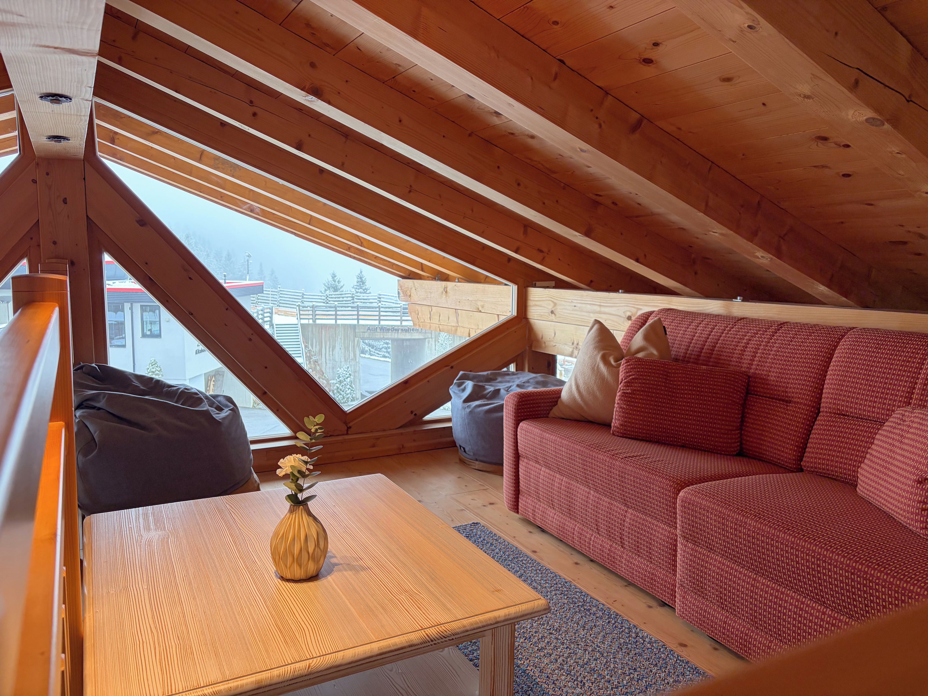 A cozy attic room with a wooden ceiling, a red sofa, a small table with a vase, and a view of a building outside.