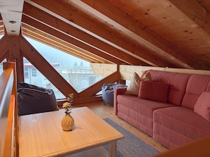 Un acogedor cuarto ático con techo de madera, un sofá rojo, una mesita con un florero y vista a un edificio.