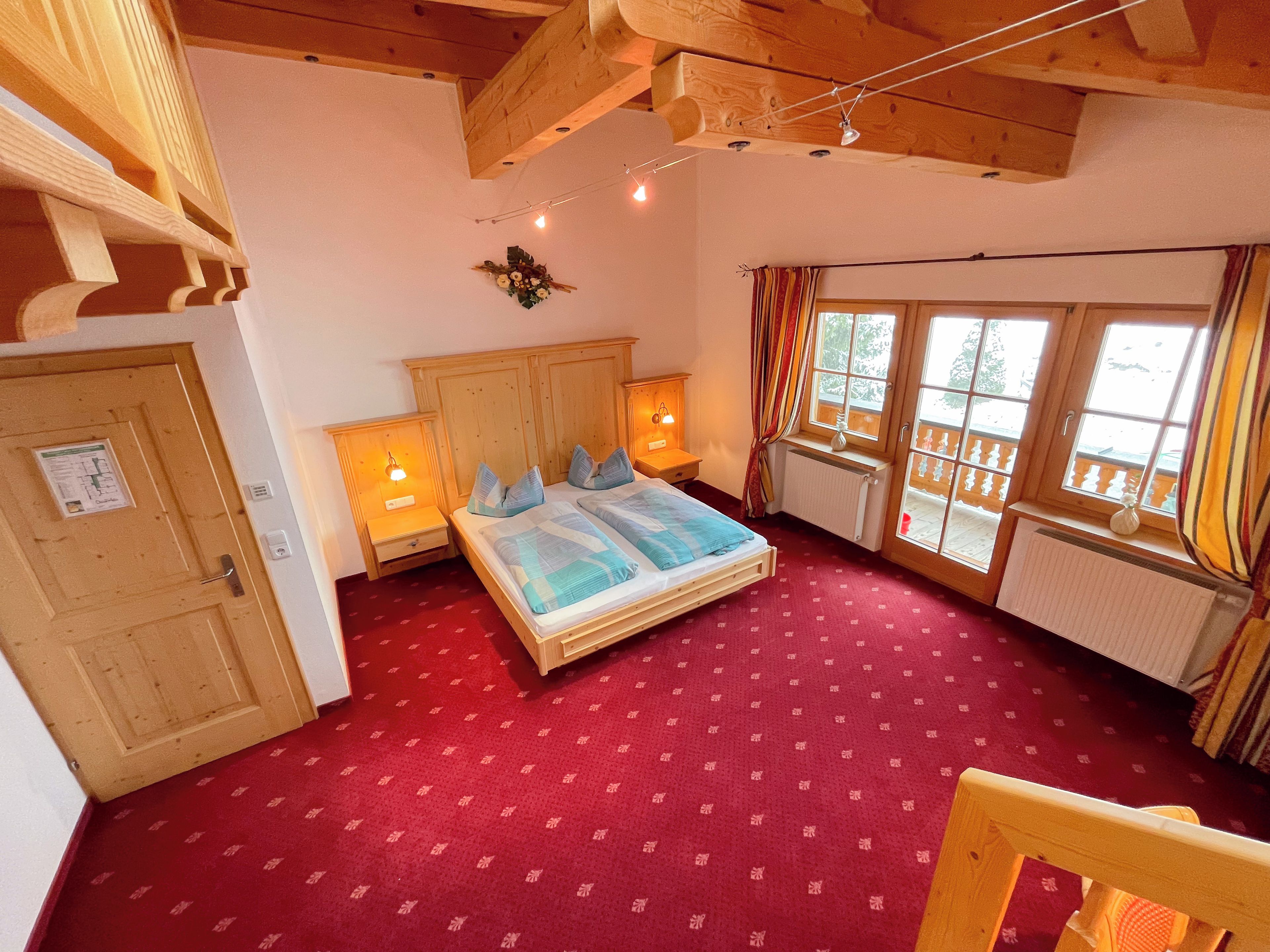 A room with a bed, wooden headboard, two bedside tables with lamps, a window with curtains, and a red carpet.