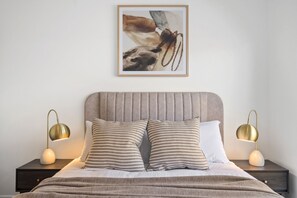 Ein Schlafzimmer mit einem Bett, zwei Nachttischlampen, einem gerahmten Kunstwerk über dem Bett und einem Nachttisch mit einer Schublade.