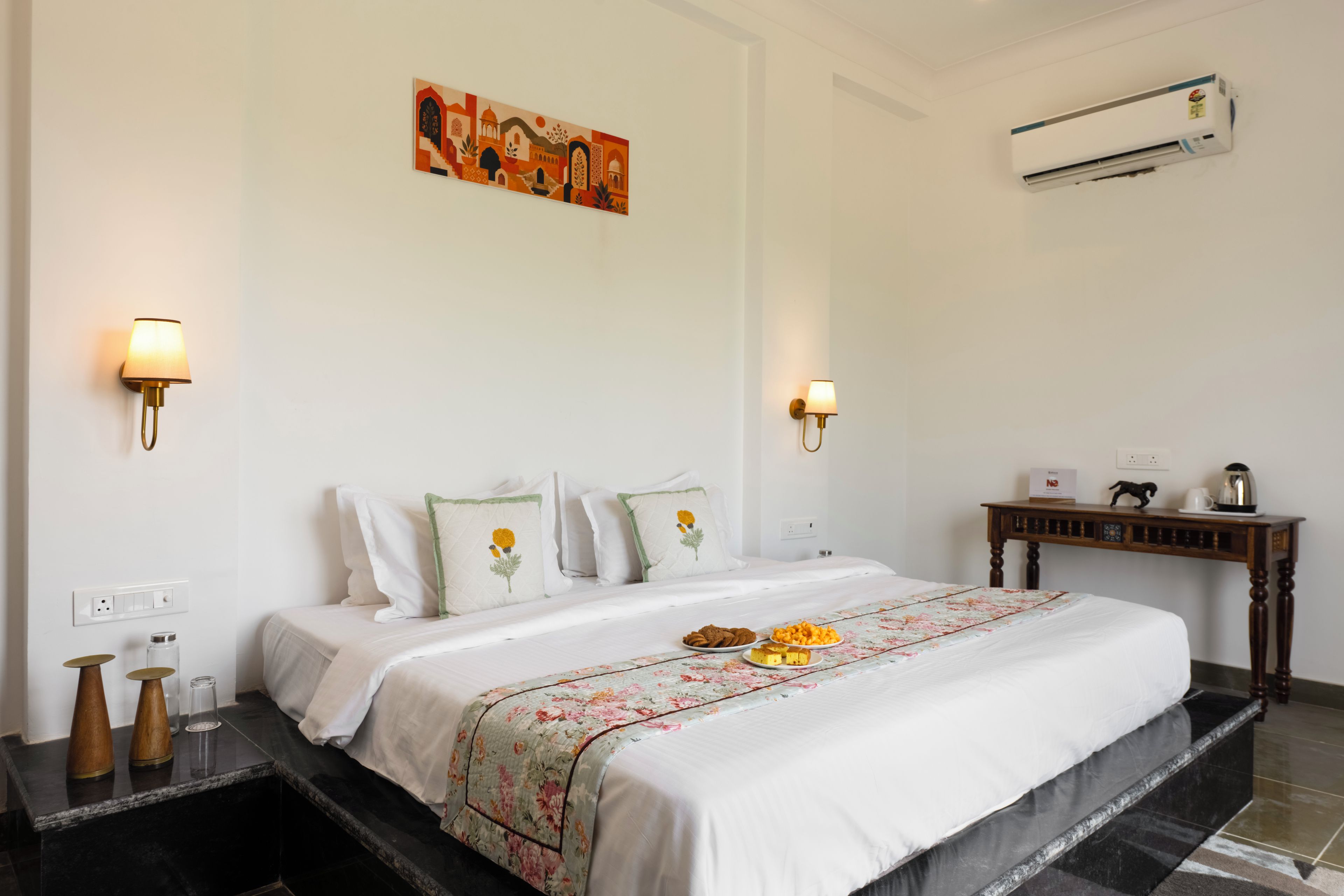A hotel room with a bed, bedside lamps, a painting, an air conditioner, a bedside table with a coffee maker, and a floral bedspread.