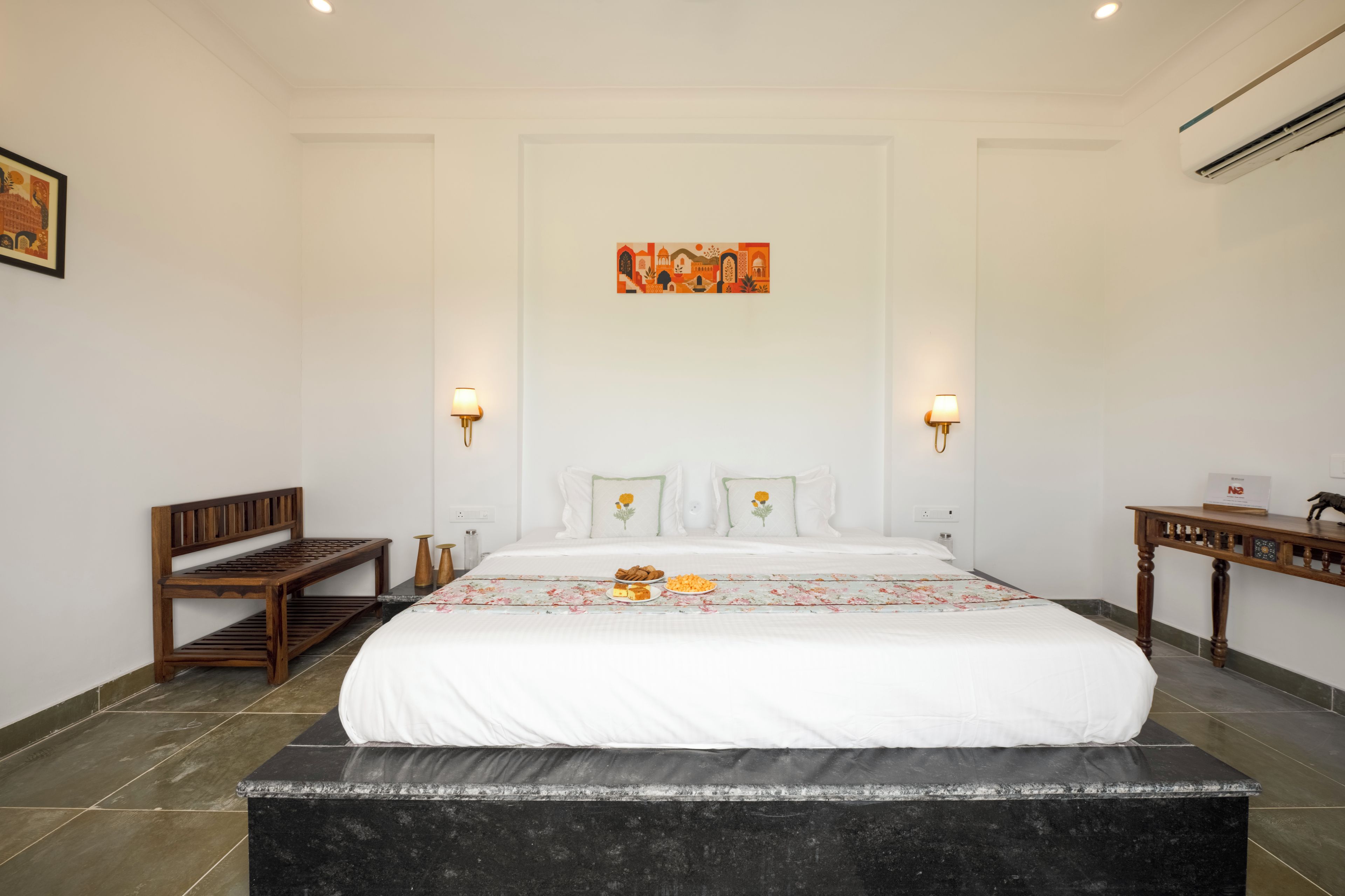 A hotel room with a large bed, a wooden bench, a small table, and a wall-mounted painting.