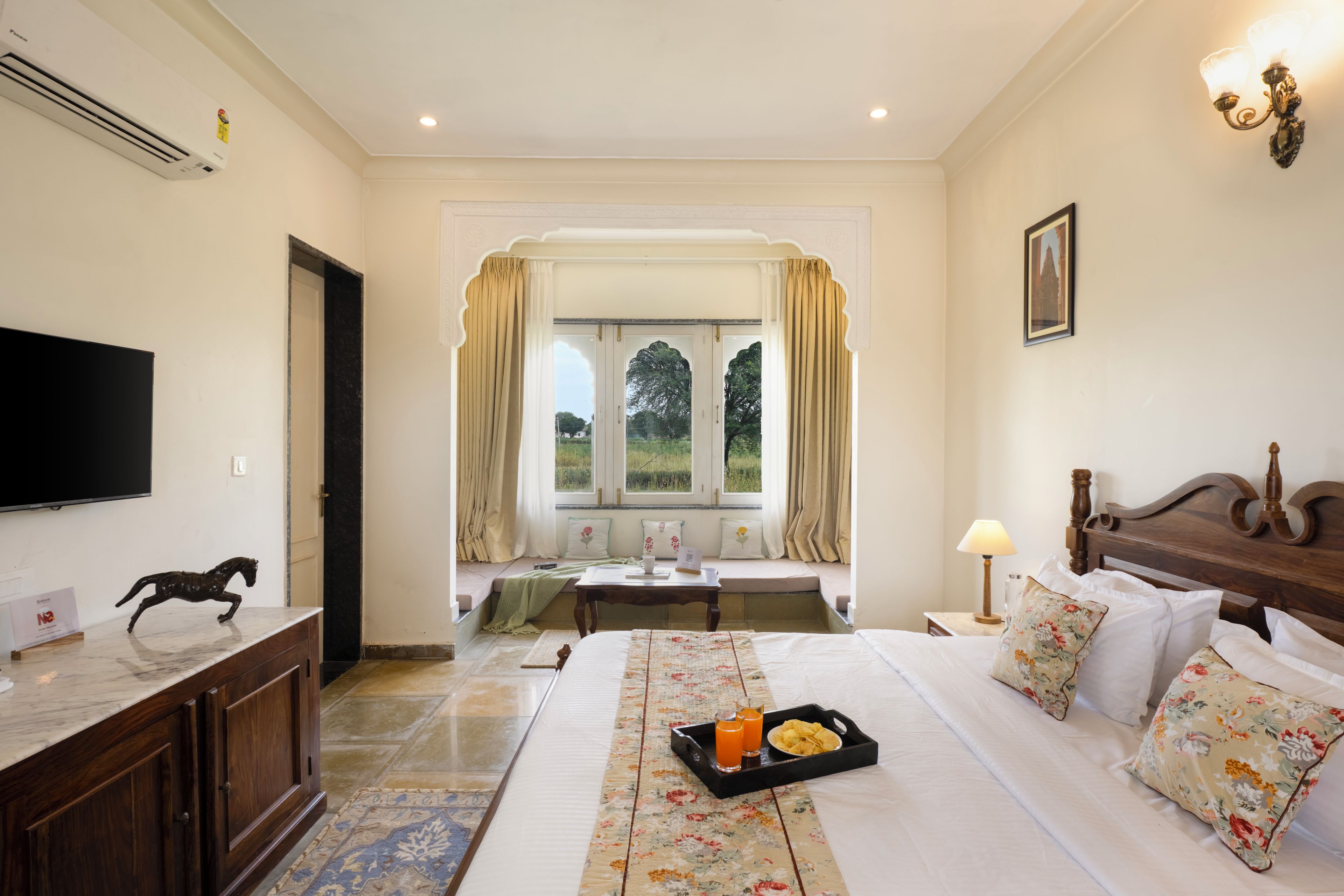 A hotel room with a bed, a television, a window with curtains, a nightstand, and a tray with snacks.