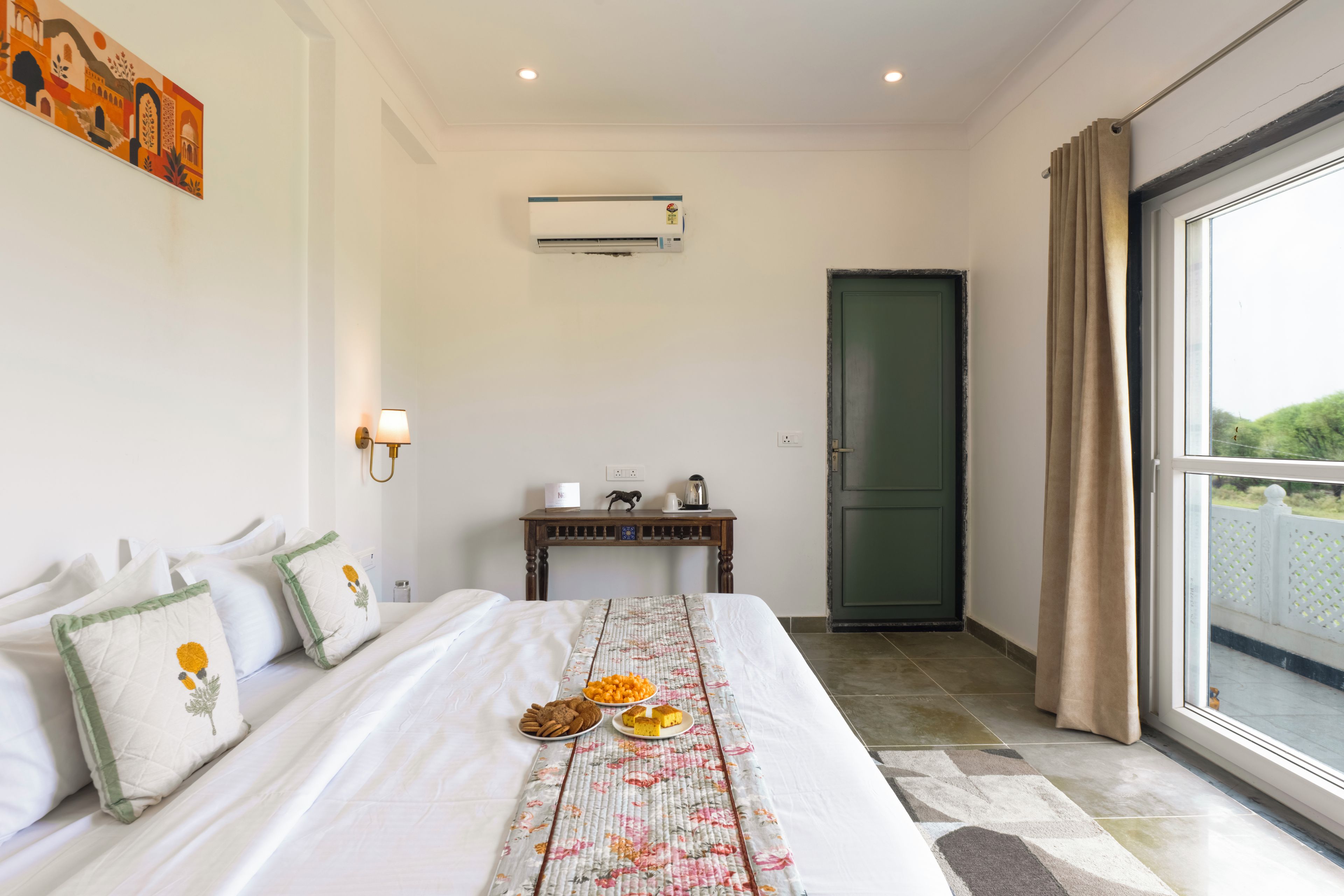 A hotel room with a bed, a table with snacks, a wall-mounted air conditioner, and a view through a window.