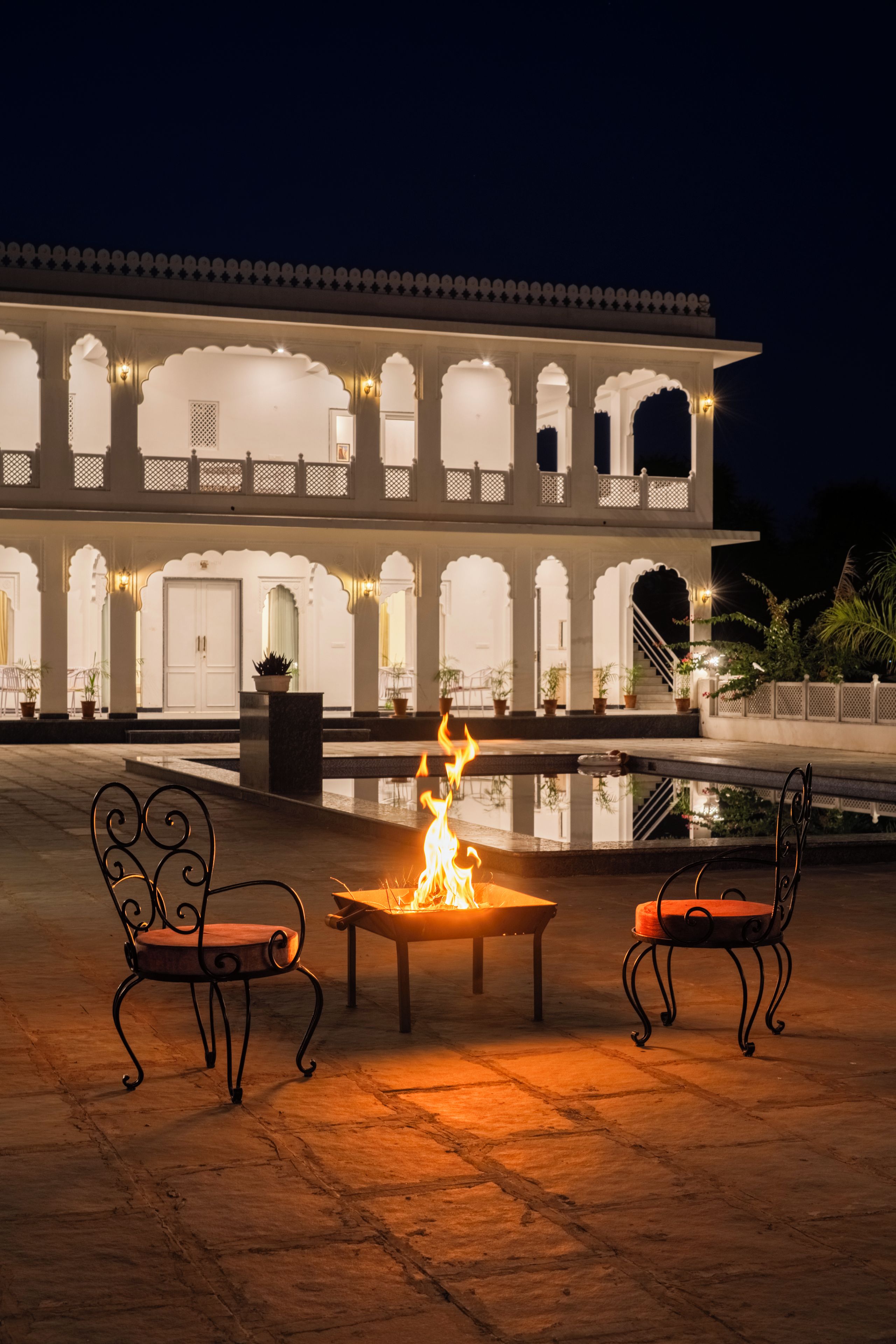 A courtyard with a fire pit and two chairs, a building with multiple arches and balconies, and a staircase leading to an upper level.