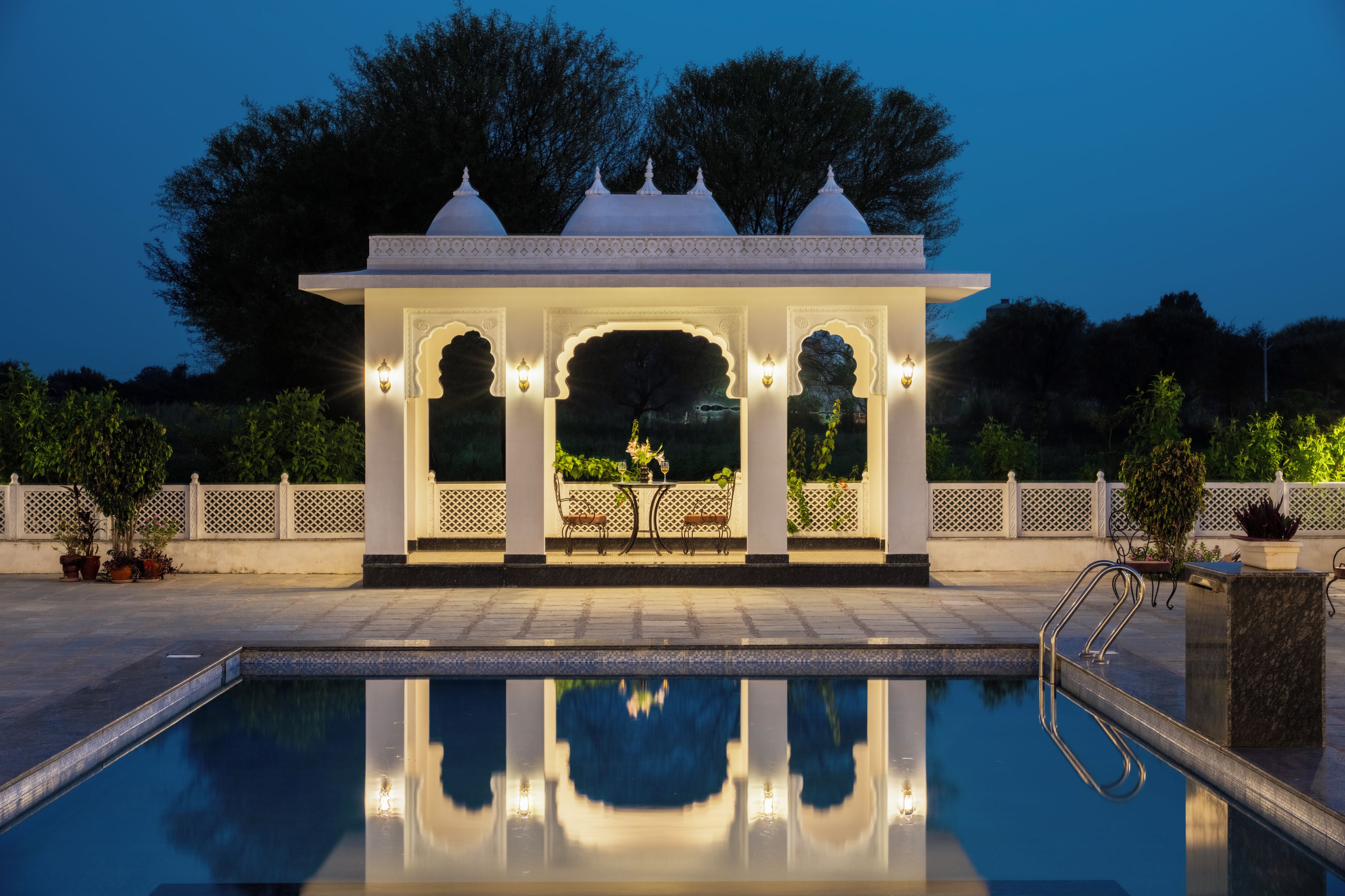 A pool with a reflection, a gazebo with arches, and a fence with plants.