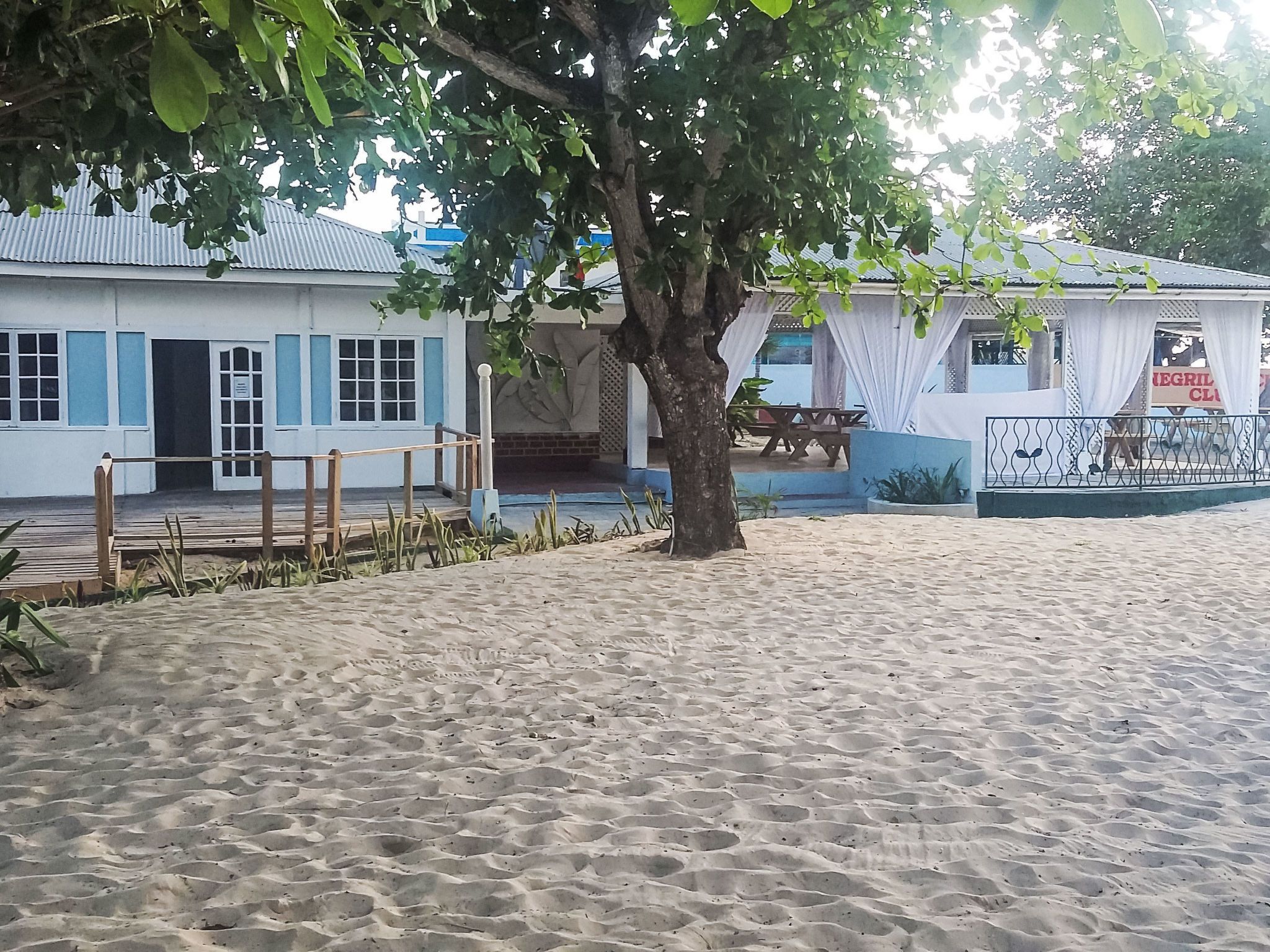 Une propriété située en bord de mer, dotée d’une plage de sable, d’une terrasse en bois, d’un bâtiment aux volets bleus et d’un arbre qui offre de l’ombre.