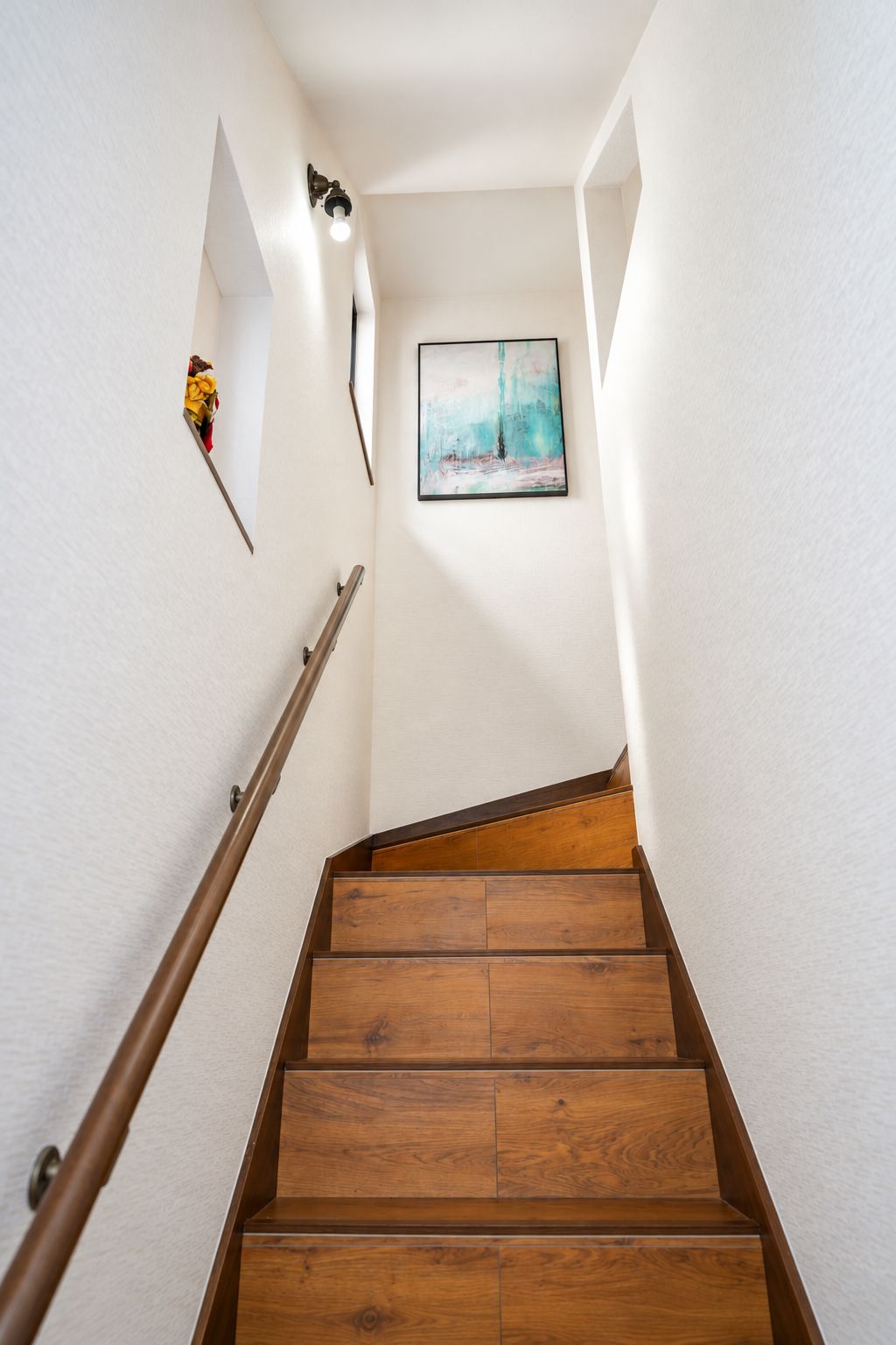 Un escalier en bois avec une rampe, des murs blancs et un tableau encadré en haut.