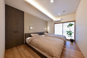 A modern bedroom with two beds, wooden flooring, a large window with blinds, and an air conditioning unit. - Hachimenzan Happo (Nakatsu)