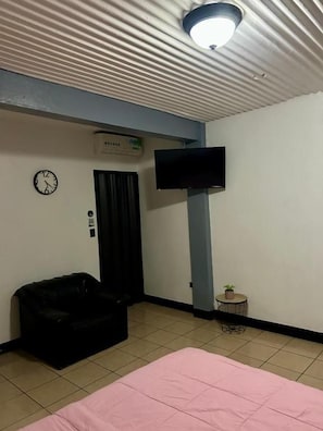 A room with a bed, a television mounted on the wall, a clock, and a small table with a plant.
