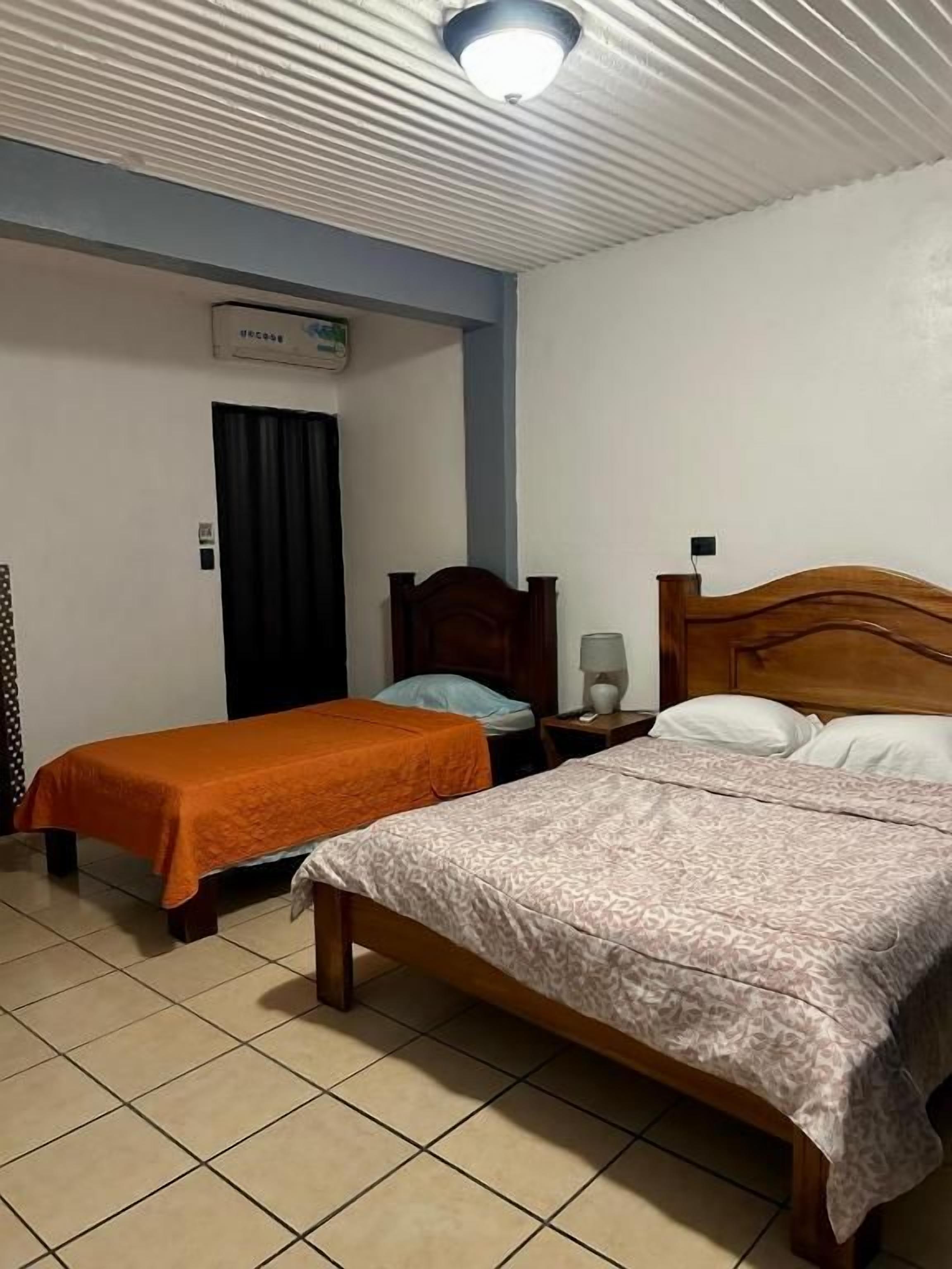 Ein Zimmer mit zwei Betten, eines mit einer gemusterten Tagesdecke und das andere mit einer orangefarbenen Decke. Ein Nachttisch mit Lampe und eine Wand-Klimaanlage sind vorhanden.