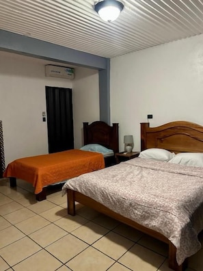 A room with two beds, one with a patterned bedspread and the other with an orange bedspread. There is a nightstand with a lamp and a wall-mounted air conditioner.