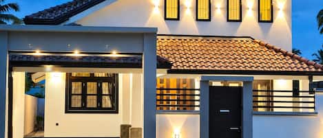 A modern two-story house with a tiled roof, a front porch with a black door, and multiple windows with exterior lighting.