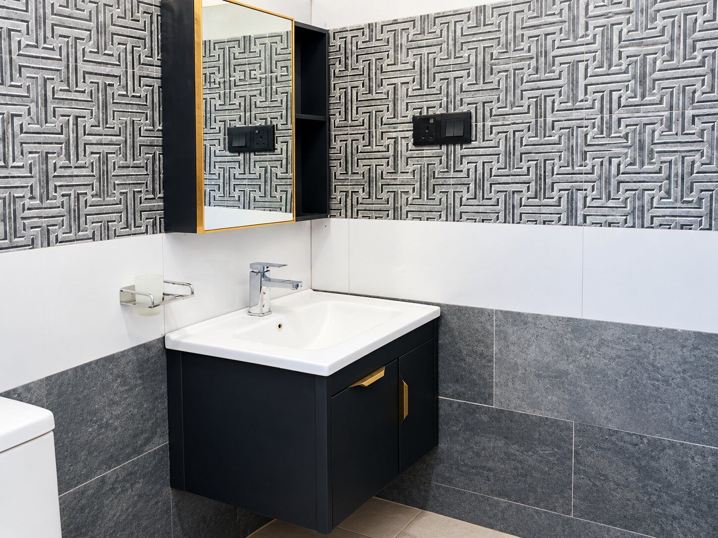 A modern bathroom with a geometric patterned wall, a white sink, and a black cabinet with gold handles.
