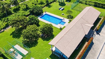 A large building with a swimming pool, surrounded by greenery and a fenced sports area.