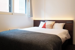 Ein Hotelzimmer mit Bett, Fenster mit Vorhängen, Nachttisch und einem an der Wand befestigten Steckdosenanschluss.