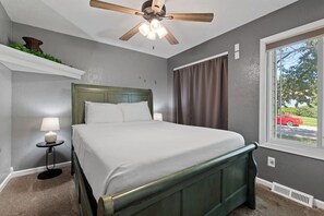 Una camera da letto con un letto, una ventola a soffitto, un comodino con una lampada, una finestra con tende e vista su un'auto rossa all'esterno.