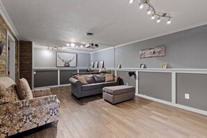 Ein modernes Wohnzimmer mit grauem Sofa, einem gemusterten Sessel, einem Hocker, Holzböden und einem Deckenprojektor.