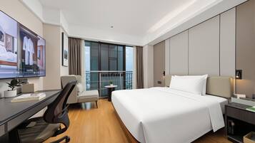 A modern hotel room with a large bed, a desk, a chair, a small table with a plant, and a view of the city through large windows.