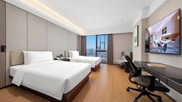 Ein Hotelzimmer mit zwei Betten, einem Schreibtisch, einem Stuhl, einem Fernseher und einem Balkon mit Stadtblick.