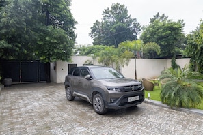 A car parked in a paved driveway with a white wall and greenery in the background. - ELIVAAS Signature DSG Farm 3BHK with Bar (New Delhi)