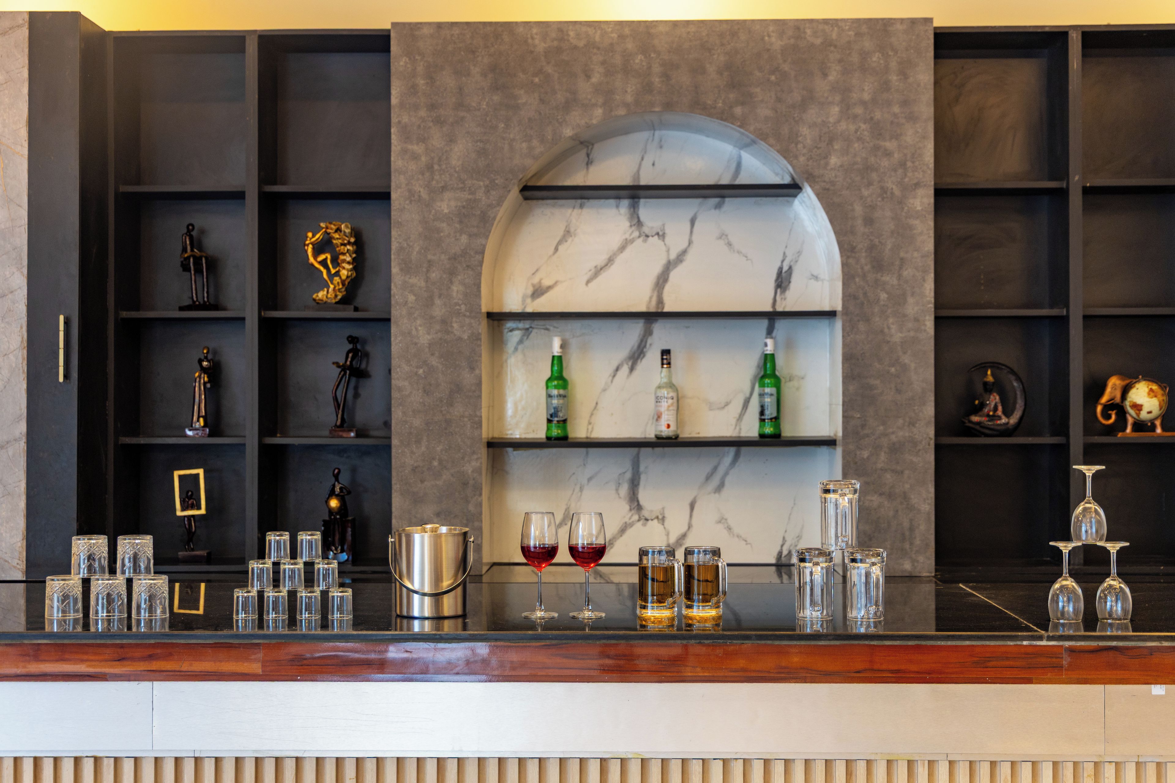 A bar area with shelves containing bottles and glasses, a marble wall with a curved arch, and a wooden counter.