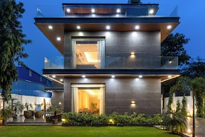 A modern multi-story building with a glass facade, outdoor seating area, and a well-lit entrance. - ELIVAAS Signature DSG Farm 3BHK with Bar (New Delhi)