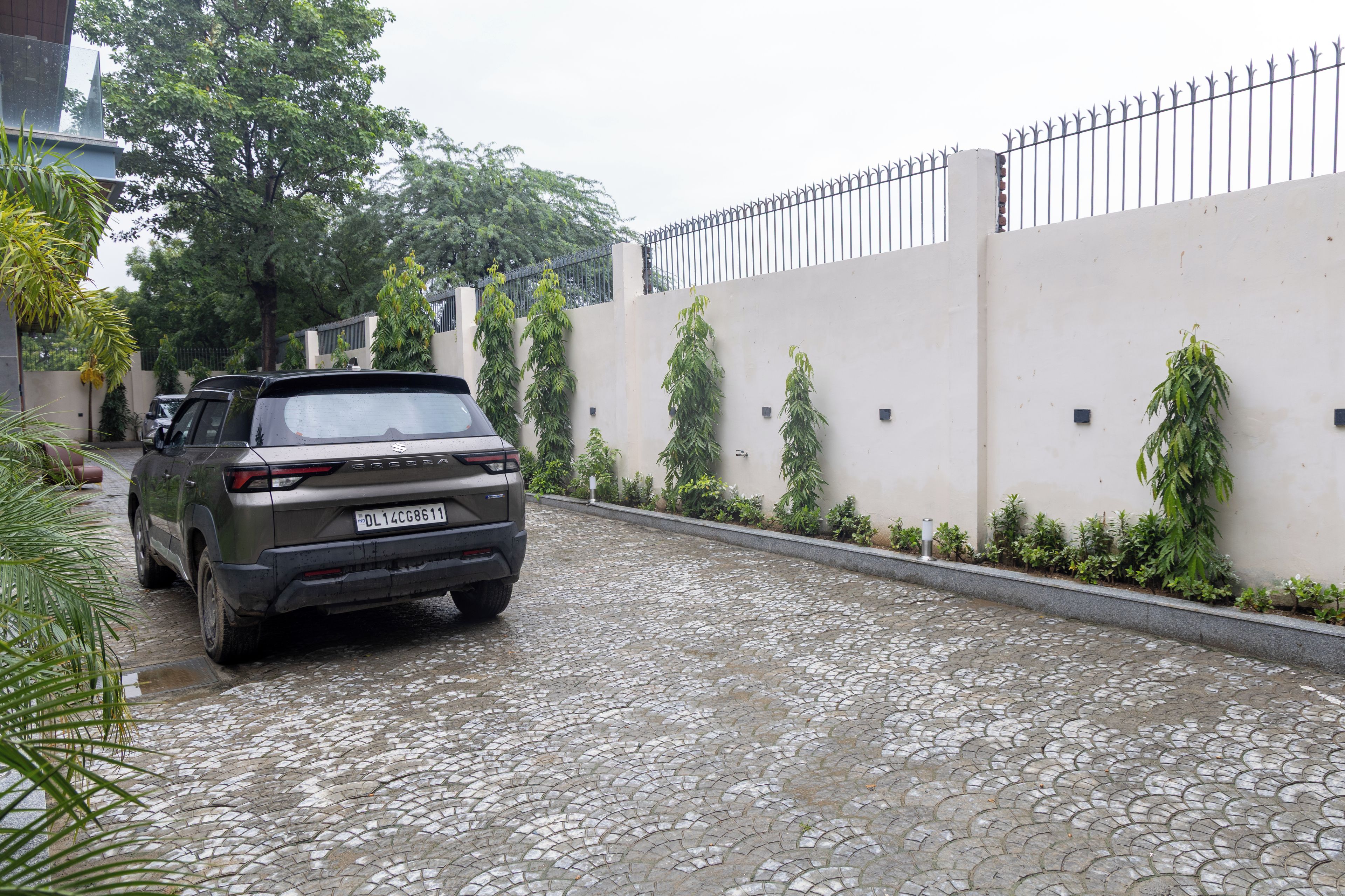 Un véhicule est garé sur une rue pavée, à côté d’un bâtiment entouré d’une clôture en métal et agrémenté de verdure.