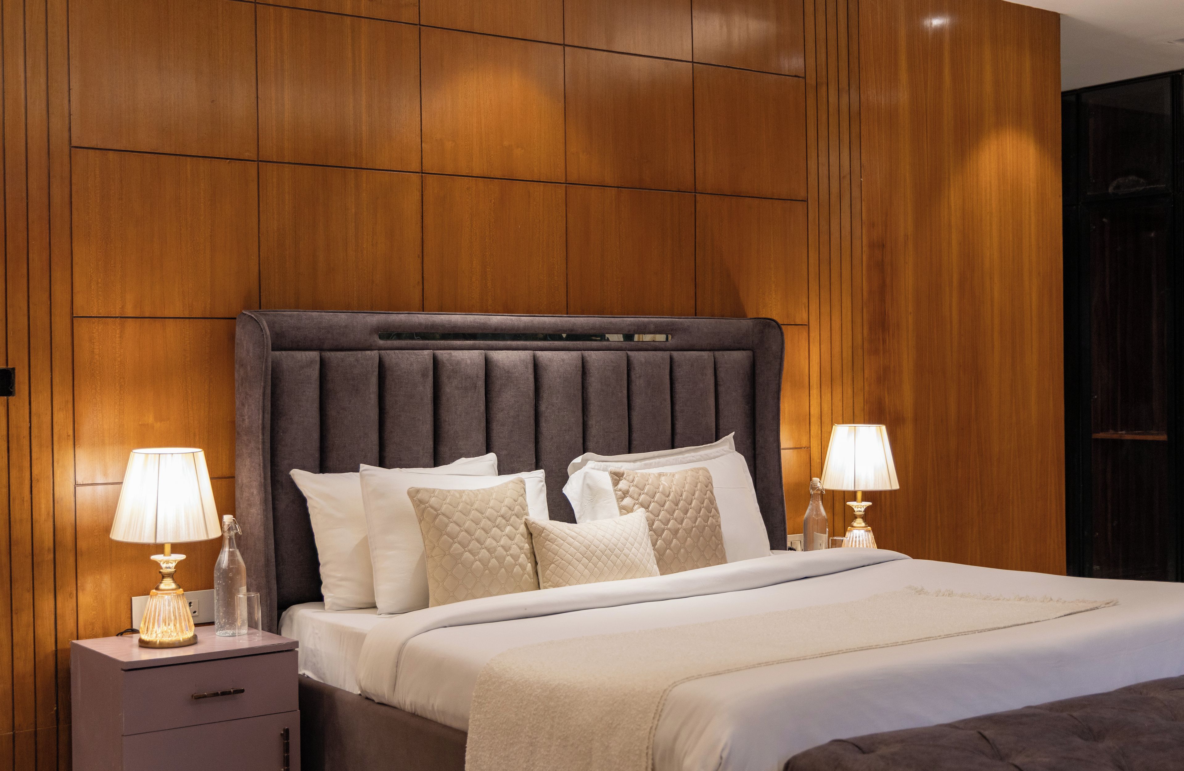 A modern bedroom with a wooden headboard, a bed with white bedding, two bedside tables with lamps, and a nightstand with a bottle and glasses.
