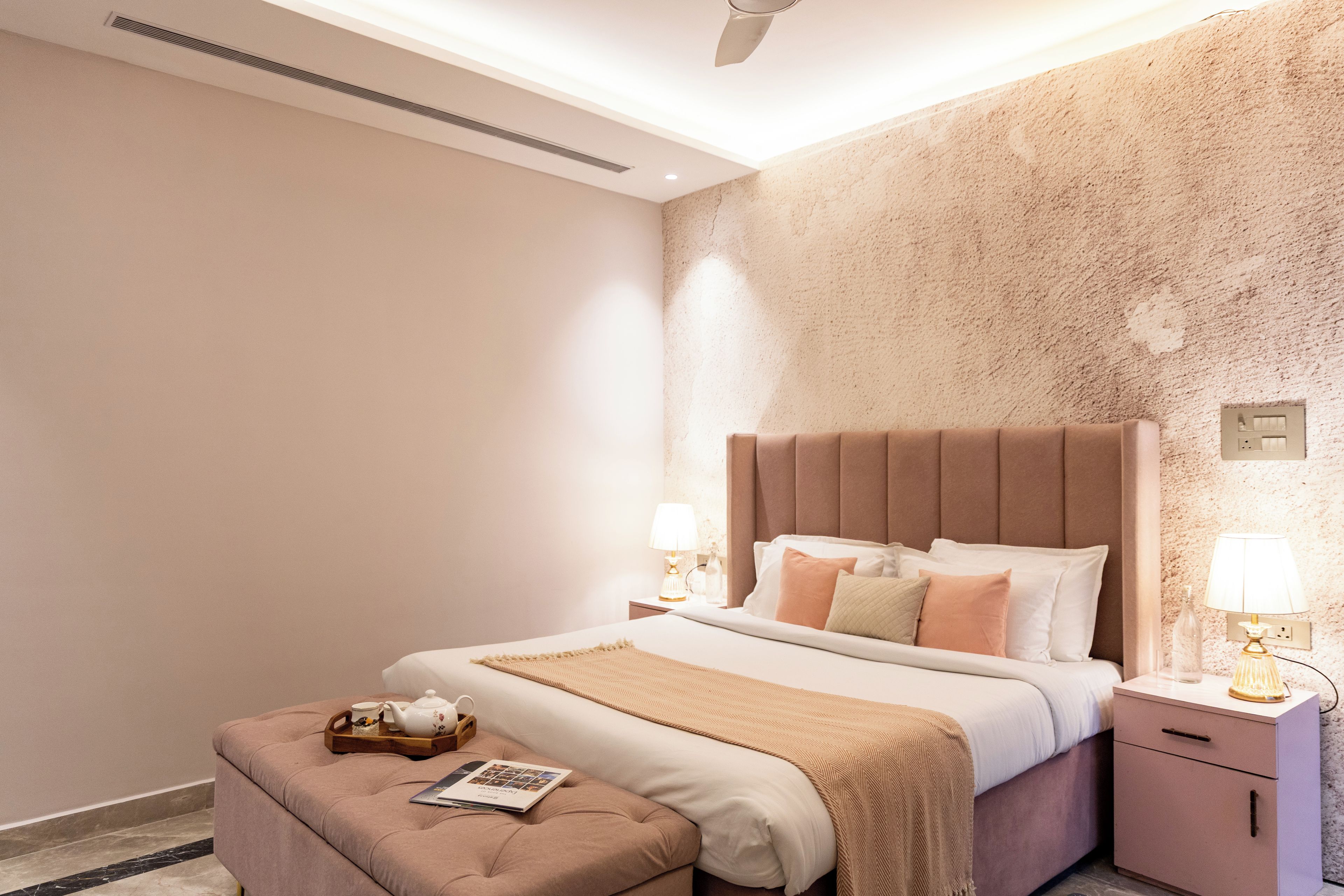 A modern bedroom with a large bed, bedside lamps, a nightstand, and a reading area with a book and teapot.