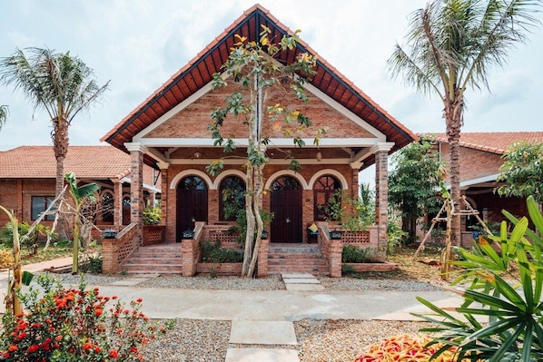 A two-story brick house with a red-tiled roof, a central entrance with a porch, and a garden with palm trees and red flowers. - DOL Phu Quoc Resort (Phu Quoc)