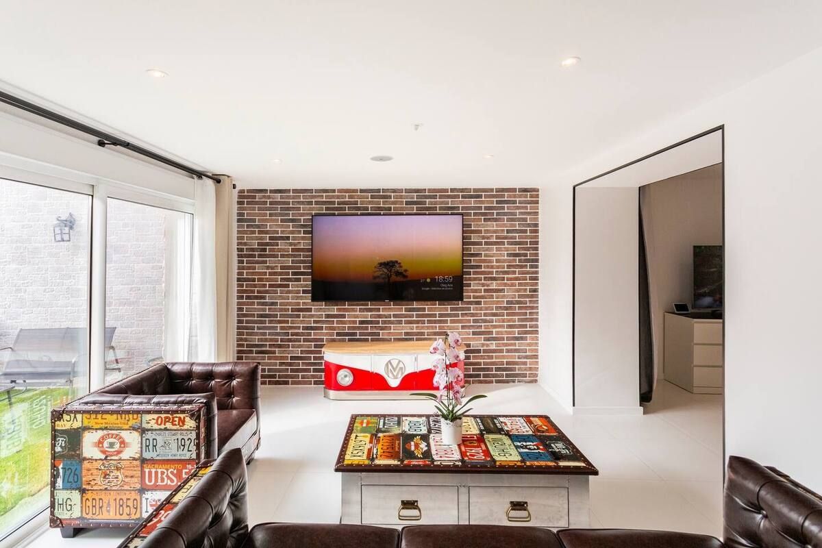 Un salón moderno con pared de ladrillo, un televisor de pantalla plana, una maleta antigua y una mesa de centro con una planta.